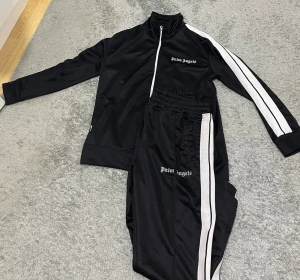 Palm Angels svart set - Svart träningsjacka från Palm Angels med vit dragkedja och vita ränder längs ärmarna. Jackan har hög krage och logotypen broderad på bröstet. Materialet är blankt och känns sportigt, perfekt för dig som gillar streetwear och snygga detaljer. Priset kan diskuteras. Ställ gärna frågor!