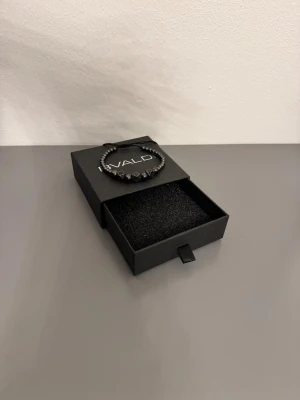 Svart armband från BVALDI - Stilrent svart armband från BVALDI med pärlor och dekorativa kubformade detaljer. Armbandet har justerbar snörning och kommer i en snygg svart ask med logga. Perfekt accessoar för dig som gillar minimalistisk och modern stil.