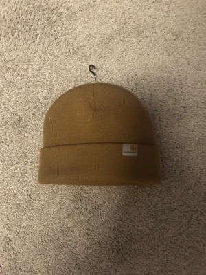 Aldrig använd brun Carhartt mössa med uppvik - Helt ny och aldrig använd snygg brun mössa från Carhartt med klassisk uppvik och logotyp framtill. Perfekt för dig som vill ha en enkel och stilren look. Tillverkad i mjukt stickat material som håller dig varm under kyliga dagar.