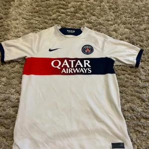 PSG vit matchtröja Nike barn L - Paris Saint-Germain fotbollströja från Nike i storlek L. Tröjan är vit med breda horisontella ränder i marinblått och rött över bröstet, PSG-logga och Nike Swoosh på framsidan. Tillverkad i Dri-Fit material som andas, med korta ärmar och ribbade detaljer vid hals och ärmslut.
