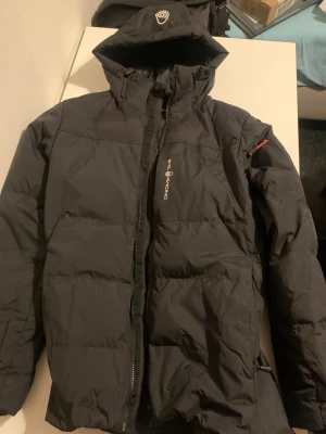 Svart pufferjacka Sail Racing - Svart pufferjacka från Sail Racing med huva och snygga detaljer som patch på ärmen och orange dragkedjedragare. Jackan har en stilren look, lång ärm och logga på bröstet. Perfekt för kalla dagar och expedition vibes.