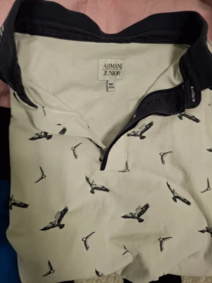 Vit piké från Armani Junior - Snygg vit piké från Armani Junior med svart krage och coolt fågelmönster över hela skjortan. Skjortan har knappar vid halsen och är tillverkad i mjuk bomull som känns skön mot huden. Perfekt för dig som gillar stilrena detaljer och unik design.