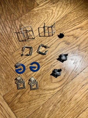 Mix av geometriska örhängen - Säljer ett coolt set med 5 par örhängen i olika former och färger. Här finns silvriga kuber, blå bokstavsörhängen, svarta och guldiga geometriska former samt kantiga hoops. Perfekt för dig som gillar statement och grafisk stil. Alla örhängen har unika detaljer och modern vibe.