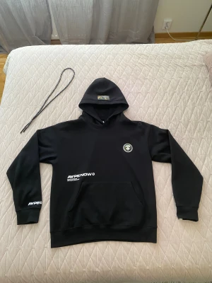 Svart hoodie från AAPE BY A BATHING APE - Svart hoodie från AAPE BY A BATHING APE med tryckt logga på bröstet och ärmen, samt stor AAPE NOW-text på ryggen. Huva med camo-detalj och tillhörande snören. Klassisk känguruficka framtill och ribbade muddar. Perfekt streetwear-vibe.