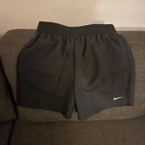 Oanvända Svarta träningsshorts från Nike - Svarta shorts från Nike med elastisk midja och standard fit. Snygg minimalistisk design med liten vit Nike-logga på benet. Perfekta för gymmet eller sport, tillverkade i lätt polyester som andas. Passar dig som gillar enkel och sportig stil. Helt oanvända, dom är medium men passar small jätte bra med.