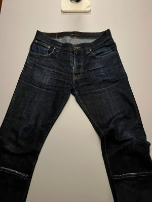 Mörkblå jeans från Nudie Jeans Co W32 L32 - Snygga mörkblå jeans från Nudie Jeans Co i klassisk femficksmodell med kontrastsömmar och broderade bakfickor. Jeansen har rak passform och är tillverkade i slitstarkt denimtyg. Perfekt för dig som gillar stilrena och tidlösa jeans.