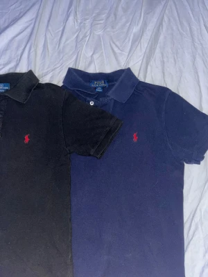 Pikétröjor från Polo Ralph Lauren - Två klassiska pikétröjor från Polo Ralph Lauren, en i marinblått och en i svart. Båda har korta ärmar, krage och knappar framtill samt den ikoniska röda logon broderad på bröstet. Perfekt för en clean och stilren look.