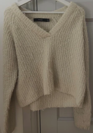 Beige stickad V-ringad tröja Vero Moda - Mysig beige stickad tröja från Vero Moda med V-ringad hals och långärmad design. Tröjan har en loose passform och grovstickad struktur som ger en chill och avslappnad vibe. Perfekt för lager-på-lager under kyliga dagar.