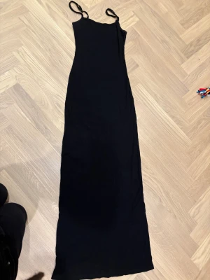 Svart långklänning från AMIS - Svart ribbad långklänning från AMIS med smala justerbara axelband. Klänningen är figurnära och har en enkel, stilren design som ger en snygg siluett. Materialet är mjukt och stretchigt, perfekt för dig som gillar minimalistisk stil.