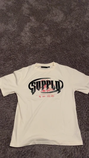 Vit Supply & Demand t-shirt med tryck - Vit t-shirt från Supply & Demand med stort svart och rött SUPPLY-tryck på bröstet och coola grafiska detaljer. Klassisk rund hals och korta ärmar. Mjuk och skön bomullskvalitet, perfekt för dig som gillar streetwear.