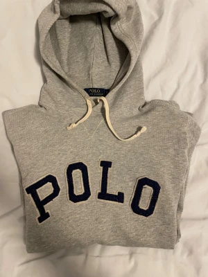 Grå hoodie från Polo Ralph Lauren - Snygg grå hoodie från Polo Ralph Lauren med stor POLO-text i mörkblått på bröstet och coolt patch med '67' vid nederkanten. Tröjan har huva med snörning, känguruficka och ribbade muddar. Perfekt för chill dagar och streetstyle.