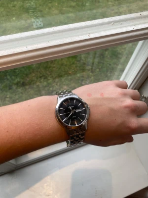 Citizen svart/silver herrklocka - Citizen herrklocka med rund svart urtavla och dag/datumvisning. Armbandet är i blankt och borstat silverfärgat metall med länkdesign. Klassisk stil med tydliga indexmarkeringar och Citizen-logga på spännet. Perfekt för dig som gillar stilrena accessoarer.