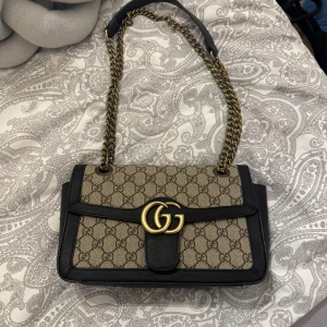 Gucci GG Marmont axelväska beige/svart - Säljer en Gucci GG Marmont axelväska i beige canvas med klassiskt GG-mönster och svarta skinnkanter. Väskan har guldfärgad kedjerem och stor GG-logo framtill. Invändigt finns dragkedjefack och ljus insida. Perfekt för dig som vill ha en ikonisk designerbag.