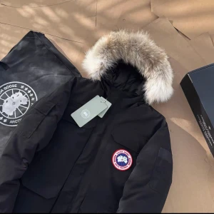 Svart Canada Goose Expedition dunjacka - Canada Goose Expedition dunjacka i svart med ikonisk patch på bröstet och stor huva med fluffig päls. Jackan har flera fickor, robust dragkedja och detaljerade logotyper. Perfekt för kalla vinterdagar och riktigt snygg streetstil. Fits S/M