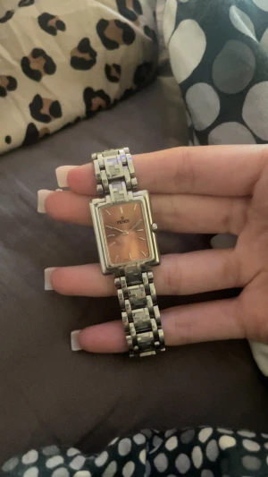 Fendi rektangulär silverklocka - Snygg Fendi klocka med rektangulärt ur och länkarmband i silverfärgat metall. Urtavlan är ljusrosa och har minimalistiska markeringar. Länken har Fendi-logga i designen och klockan har en stilren vibe som passar till många outfits osäker på storleken .