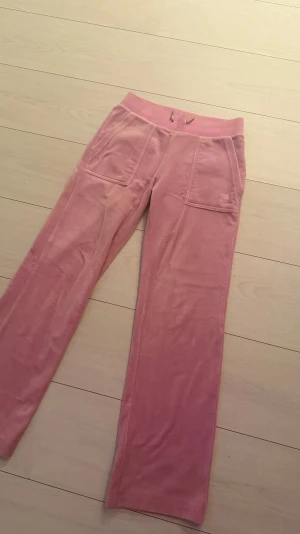 Rosa velourbyxor Juicy Couture XS - Säljer ett par rosa mjukisbyxor från Juicy Couture i storlek XS. Byxorna har bred resårmidja med snörning, stora fickor fram och bak med metallknappar, samt broderad JC-logga på ena fickan. Materialet är mjuk velour som ger en skön och lyxig känsla. Nypris 1200kr