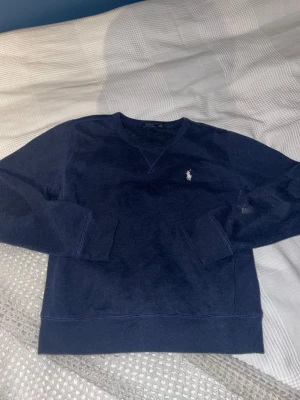 Mörkblå tröja från Polo Ralph Lauren - Mörkblå crewneck från Polo Ralph Lauren med klassisk vit broderad logga på bröstet. Tröjan har rund halsringning, långa ärmar och ribbade muddar. Skön och stilren modell i mjuk bomull, perfekt till jeans eller chinos.