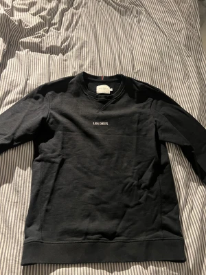 Svart sweatshirt från Les Deux, M pris går att diskutera  - Svart sweatshirt från Les Deux med stilren vit logotyp broderad på bröstet. Tröjan har rund hals, ribbade muddar och lång ärm. Materialet är mjuk bomull som känns skönt mot huden. Perfekt för dig som gillar minimalistisk och clean stil. Passar även folk som har storlek .