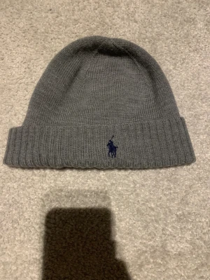Grå merinoullsmössa från Polo Ralph Lauren - Snygg grå mössa från Polo Ralph Lauren i mjuk och tvättbar merinoull. Klassisk ribbstickad design med uppvikt kant och mörkblå broderad Polo-logga framtill. Perfekt för kalla dagar och enkel att matcha med din stil.
