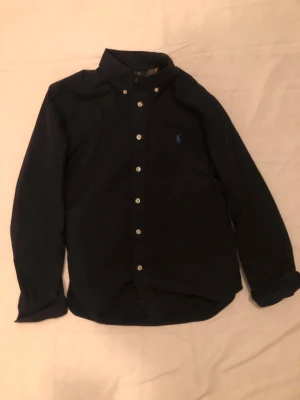 Svart skjorta från Polo Ralph Lauren - Svart skjorta från Polo Ralph Lauren med klassisk krage och knappar framtill, nypris är 1160. Endast använd ett fåtal gånger. Om du har några funderingar så kan du ta och kontakta mig så svarar jag genast😊