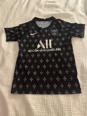 PARIS SAINT-GERMAIN TRÄNINGS T-SHIRT PRE MATCH - SVART/VIT/ BARN - Snygg svart PSG fotbollströja från Nike med vitt tryck och logga. Tröjan har korta ärmar och är täckt av ett unikt mönster med siffror. Materialet är lätt och andas, perfekt för träning eller match. Paris Saint-Germain-emblem på bröstet och sponsortryck fram och bak.