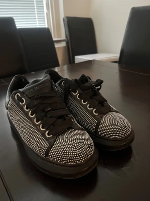 Steve Madden svarta sneakers med nitar - Svarta sneakers från Steve Madden med glittriga silverfärgade nitar över hela skon. Klassisk rund tå, snörning och platt sula. Skorna har en cool och edgy vibe, perfekta för att sticka ut. Materialet är syntet med detaljer i skinnimitation.
