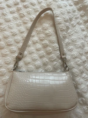 Beige axelväska med krokopräglat mönster - Snygg beige axelväska med krokopräglat mönster och silverfärgade detaljer. Väskan har en avtagbar axelrem och stängs med dragkedja upptill. Perfekt storlek för det viktigaste och ger en clean vibe till din outfit.