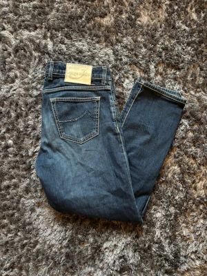 Jacob Cohen - Snygga blå jeans från Jacob Cohen med klassisk femficksdesign och kontrastsömmar. Jeansen har knäppning med synliga metallknappar och en beige patch bak i midjan. Materialet är mjukt jeans-tyg som sitter skönt och har en slim passform.