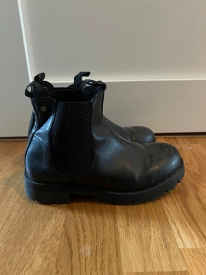 Svarta chelsea boots från Linear 35 - Svarta chelsea boots från Linear of Sweden med grov platt sula och elastiska paneler på sidorna. Tillverkade i skinn med rund tå och dragflikar fram och bak. Storlek 35 