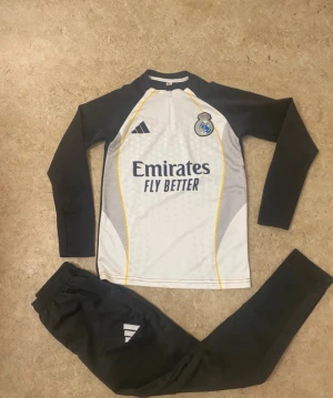 Real Madrid långärmad fotbollströja Adidas - Säljer en långärmad Real Madrid fotbollströja från Adidas i svart och vitt med gula detaljer. Tröjan har dragkedja vid halsen, klubbmärke och sponsortryck på bröstet. Materialet är lätt och andas, perfekt för träning eller match.
