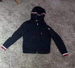 Moncler kofta - Snygg och häftig zip Hoodie från Moncler. Tröjan är mörkblå med vit röda ränder på luvan respektive armmuddar. Tröjan är både extremt snygg men även bekväm vilket gör det perfekt för en chill men utstickande outfit! Koftan är i strl S. Skick: 9/10💯Skriv vid minsta fundering!