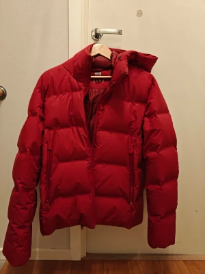 Röd pufferjacka från Uniqlo - Riktigt fet röd pufferjacka från Uniqlo med huva och dragkedja. Jackan är vadderad och har två fickor med dragkedja framtill. Perfekt för kalla dagar när du vill hålla stilen och värmen. Klassisk passform och snygg, enkel design.
