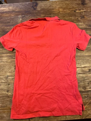 Röd pikétröja från Polo Ralph Lauren - Snygg röd pikétröja från Polo Ralph Lauren med klassisk krage och korta ärmar. Tröjan har den ikoniska blå Polo-loggan broderad på bröstet och är gjord i mjuk bomull. Perfekt för dig som gillar stilrena plagg.