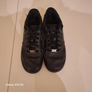 Nike Air Force 1 LE helsvarta sneakers - Säljer ett par klassiska Nike Air Force 1 LE sneakers i helsvart skinn. Modellen har låg profil, rund tå, snörning och den ikoniska swoosh-loggan på sidan. Perfekta för dig som gillar en clean och tidlös look.