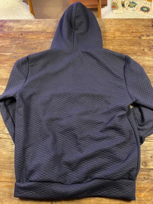 Mörkblå hoodie med dragkedja - Säljer en mörkblå hoodie med snyggt quiltat mönster och dragkedja framtill. Tröjan har huva med snörning och långa ärmar. Okänt märke. Stor i storlek