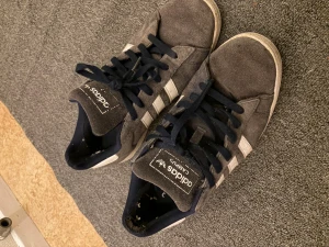 Adidas Campus 00 sneakers  - Säljer ett par klassiska Adidas Campus 00 sneakers med vita detaljer och de ikoniska tre ränderna på sidorna. Skorna har svart snörning, vit hälkappa med Adidas-logga och platt sula. Perfekta för en streetwear-look.