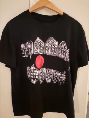 Svart t-shirt från Pixiesleeze - Svart t-shirt från Pixiesleeze med stor grafisk print av tänder och en röd smiley på framsidan. Baksidan har ett stort tryck med märkesnamnet i vit, edgy stil. Modellen är klassisk med rund hals och korta ärmar, perfekt för dig som gillar streetwear och unika motiv.