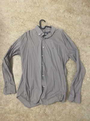 Grå skjorta från Tommy Hilfiger - Snygg grå skjorta från Tommy Hilfiger med klassisk krage och diskret logga på bröstet. Skjortan har lång ärm, vita knappar och är tillverkad i mjuk bomull. Perfekt för dig som gillar stilrena och enkla plagg med en touch av premiumkänsla.