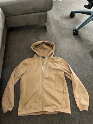 Beige hoodiejacka från Sail Racing stl 160 - Beige hoodiejacka från Sail Racing med dragkedja framtill och en extra bröstficka med zip. Jackan har broderad logga på bröstet, huva och långa ärmar. Insidan är mjuk och fleecefodrad för extra komfort. Perfekt för chill dagar och streetwear.