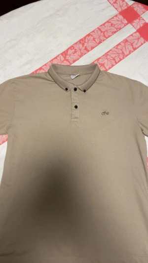 Beige pikétröja från Lacoste M - Snygg beige pikétröja från Lacoste i klassisk modell med krage och tre knappar framtill. Tröjan har det ikoniska krokodilmärket broderat på bröstet och är tillverkad i bomull. Perfekt för en clean och stilren look.