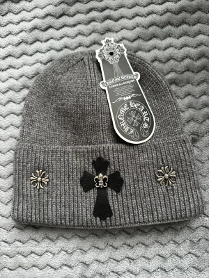 Grå Chrome Hearts x Comme des Garçons mössa - Snygg grå ribbstickad mössa med coola silvriga metallblommor och svart läderpatch med korsdetalj framtill. Märkt med Chrome Hearts x Comme des Garçons tag. Aldrig använd. Skriv om du har några frågor 