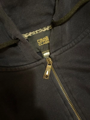 Mörkblå  hoodiejacka från Cavalli Class - Mörk blå hoodiejacka från Cavalli Class med dragkedja i guld och coola logodetaljer längs ärmarna. Jackan har huva med snörning, ribbade muddar och fickor fram. Materialet är mjuk bomull och trycket på bröstet ger extra stilpoäng.