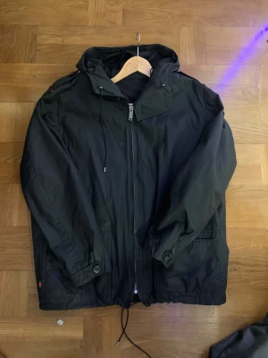 Svart jacka från Gucci - Svart Gucci jacka med huva och dragkedja framtill. Jackan har snörning nedtill, knappdetaljer vid ärmslut och fickor, samt klassisk Gucci-rand på axeln. Materialet är syntet och jackan har en loose passform, perfekt regn men även snö. Priser går alltid att diskuteras. 