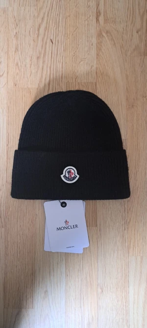 Svart Moncler mössa i ull - Svart ribbstickad mössa från Moncler med klassisk uppvikt kant och broderad Moncler-logga framtill. Tillverkad i mjuk ull som håller dig varm och snygg under kyliga dagar. Perfekt accessoar för dig som gillar stilrena och exklusiva detaljer.