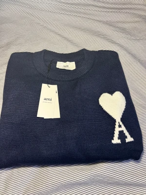 Mörkblå stickad tröja från Ami - Snygg mörkblå stickad tröja från Ami Alexandre Mattiussi med ett stort vitt hjärta och bokstaven A på bröstet. Tröjan har rund hals, ribbade muddar och lång ärm. Perfekt för dig som gillar stilrena detaljer och vill ha något som sticker ut. 0bs lnte 0rglnal‼️
