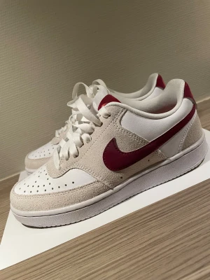 Nike sneakers i vitt och vinrött - Nike sneakers med vit och beige mocka samt vinröda detaljer och swoosh. Låg modell med snörning, perforerad tå och platt sula. Insidan har vinröd innersula med Nike-logga. Perfekt för dig som gillar stilrena och sportiga sneakers.
