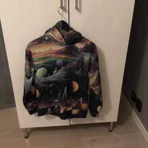 Galaxy hoodie med färgglatt rymdmotiv - Unik hoodie med ett färgglatt rymdmotiv som visar planeter, berg och galaxer i olika nyanser av lila, blått, gult och svart. Tröjan har huva och långa ärmar, tillverkad i mjukt syntetmaterial. Perfekt för dig som gillar coola och fantasifulla prints.