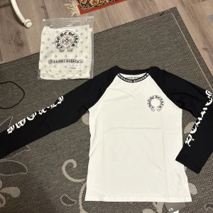 Chrome Hearts svartvit långärmad tröja - Säljer en svartvit långärmad tröja från Chrome Hearts med kontrastärmar och stora tryck på både bröst, rygg och ärmar. Tröjan har klassisk rund hals och coola gotiska logotyper. Tillverkad i mjuk bomull, perfekt för dig som gillar streetwear vibes.