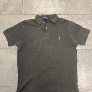 Mörkgrå pikétröja från Ralph Lauren - Snygg mörkgrå pikétröja från Polo Ralph Lauren i bomull. Klassisk krage, två knappar och väldigt fin passform