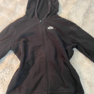 Svart Nike hoodie med dragkedja - Svart hoodie från Nike med vit logga på bröstet. Tröjan har huva, hel dragkedja framtill och två stora fickor. Standard fit och tillverkad i mjukt material som passar perfekt till chill eller träning.